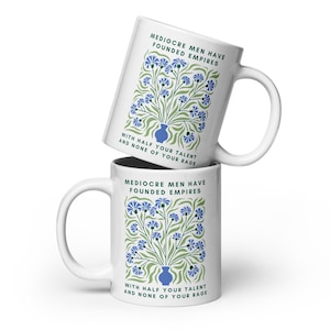 Puede incluir: Dos tazas de cerámica blanca apiladas. Cada taza presenta un diseño floral con flores azules y tallos verdes, y el texto "MEDIOCRE MEN HAVE FOUNDED EMPIRES" y "WITH HALF YOUR TALENT AND NONE OF YOUR RAGE."