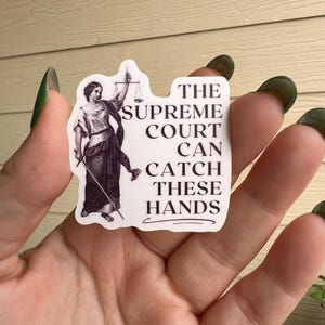 Op de afbeelding: Een witte sticker met een zwart-witte illustratie van een vrouw die een weegschaal en een zwaard vasthoudt. De tekst op de sticker luidt "The Supreme Court Can Catch These Hands."