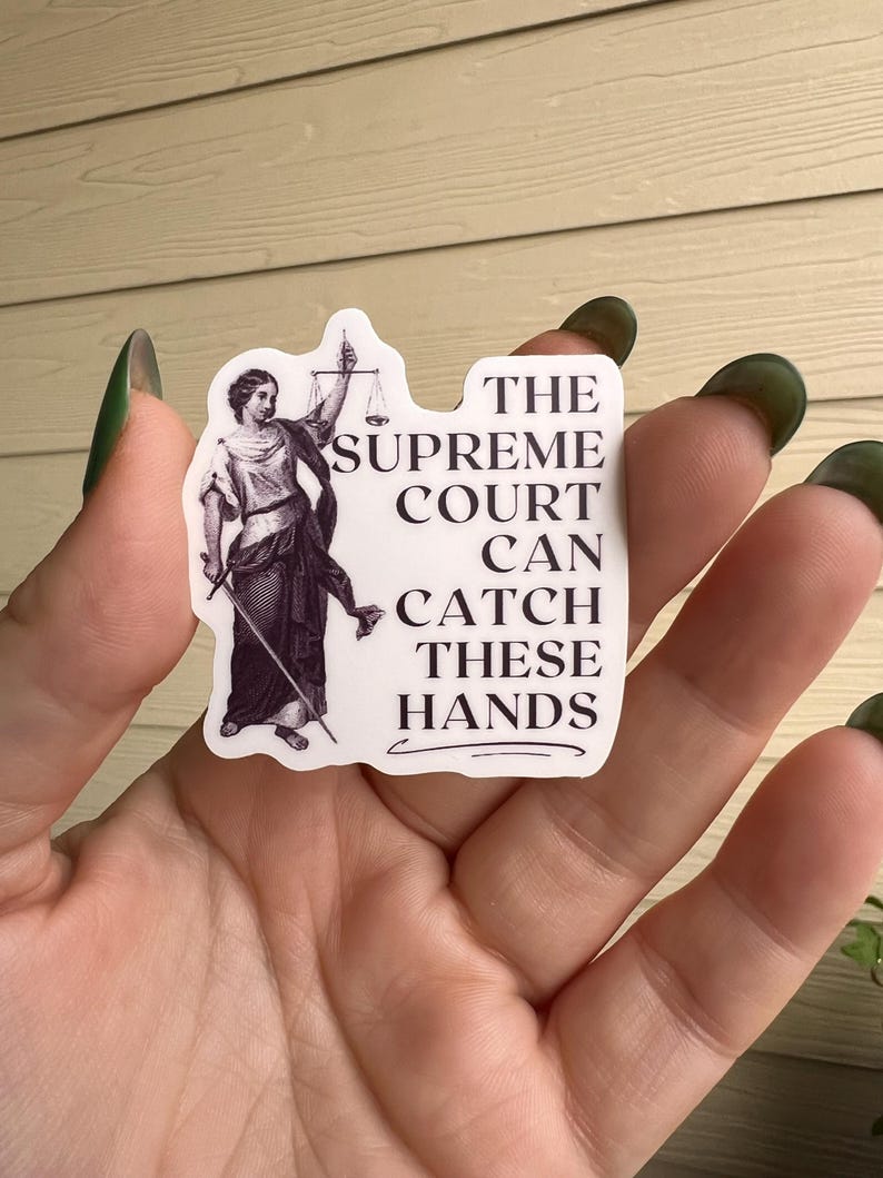 Op de afbeelding: Een witte sticker met een zwart-witte illustratie van een vrouw die een weegschaal en een zwaard vasthoudt. De tekst op de sticker luidt "The Supreme Court Can Catch These Hands."
