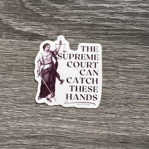 Op de afbeelding: Een witte sticker met een zwart-witte illustratie van een vrouw die een weegschaal en een zwaard vasthoudt. De tekst op de sticker luidt "The Supreme Court Can Catch These Hands."