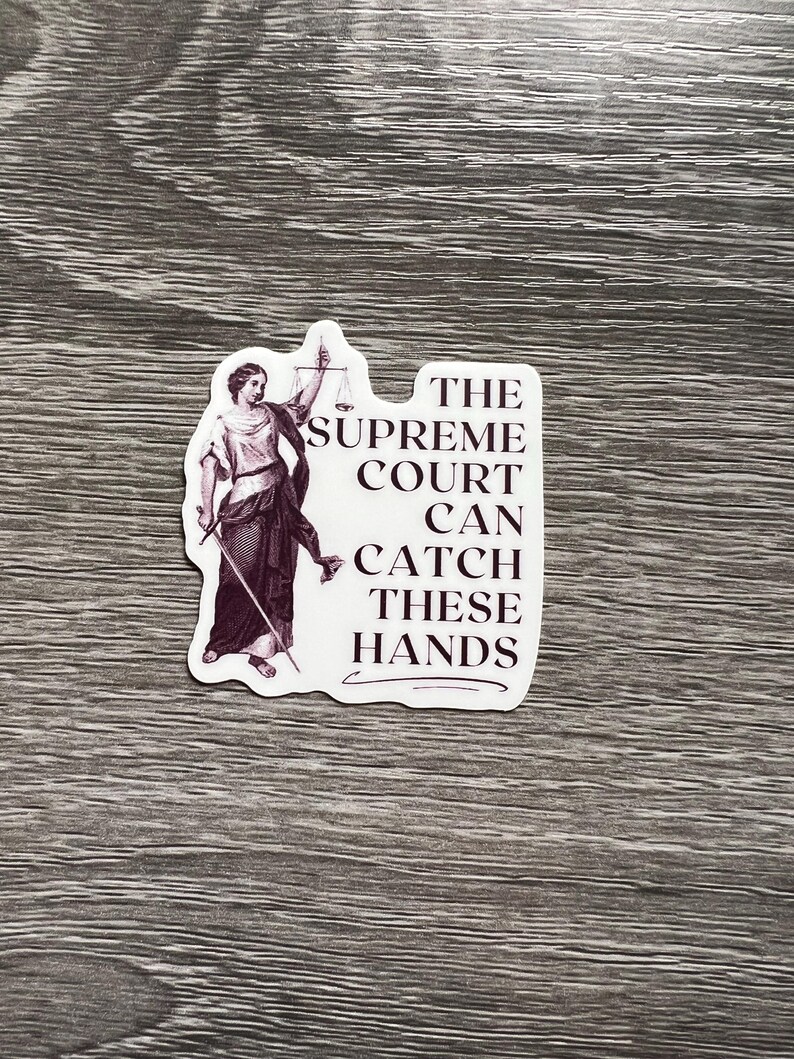 Op de afbeelding: Een witte sticker met een zwart-witte illustratie van een vrouw die een weegschaal en een zwaard vasthoudt. De tekst op de sticker luidt "The Supreme Court Can Catch These Hands."