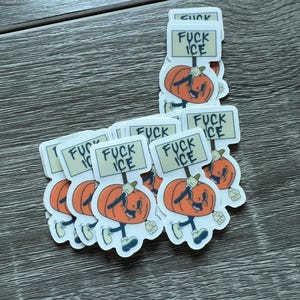 May include: A stack of stickers featuring a cartoon pumpkin character holding a sign that reads "FUCK ICE". The pumpkin is orange with a smiling face, blue shoes, and white gloves. The background is a wooden surface.