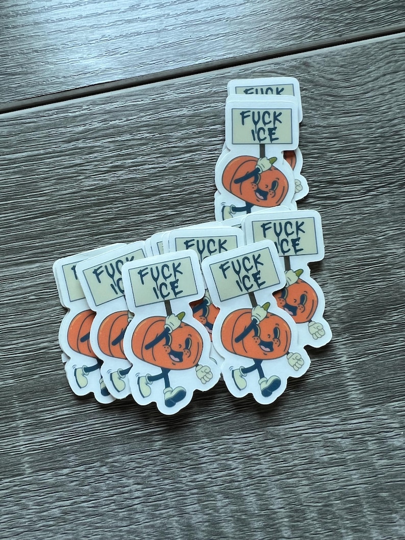 May include: A stack of stickers featuring a cartoon pumpkin character holding a sign that reads "FUCK ICE". The pumpkin is orange with a smiling face, blue shoes, and white gloves. The background is a wooden surface.