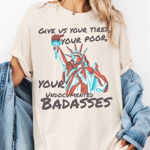 May include: Cream-colored t-shirt featuring a graphic of the Statue of Liberty in red and blue, holding a torch. The shirt has the text "Give us your tired, your poor, your undocumented badasses."