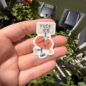 May include: A sticker featuring a cartoon pumpkin character holding a sign that reads "FUCK ICE". The pumpkin is orange with a smiling face, blue shoes, and a white glove. The sticker is held in a hand.