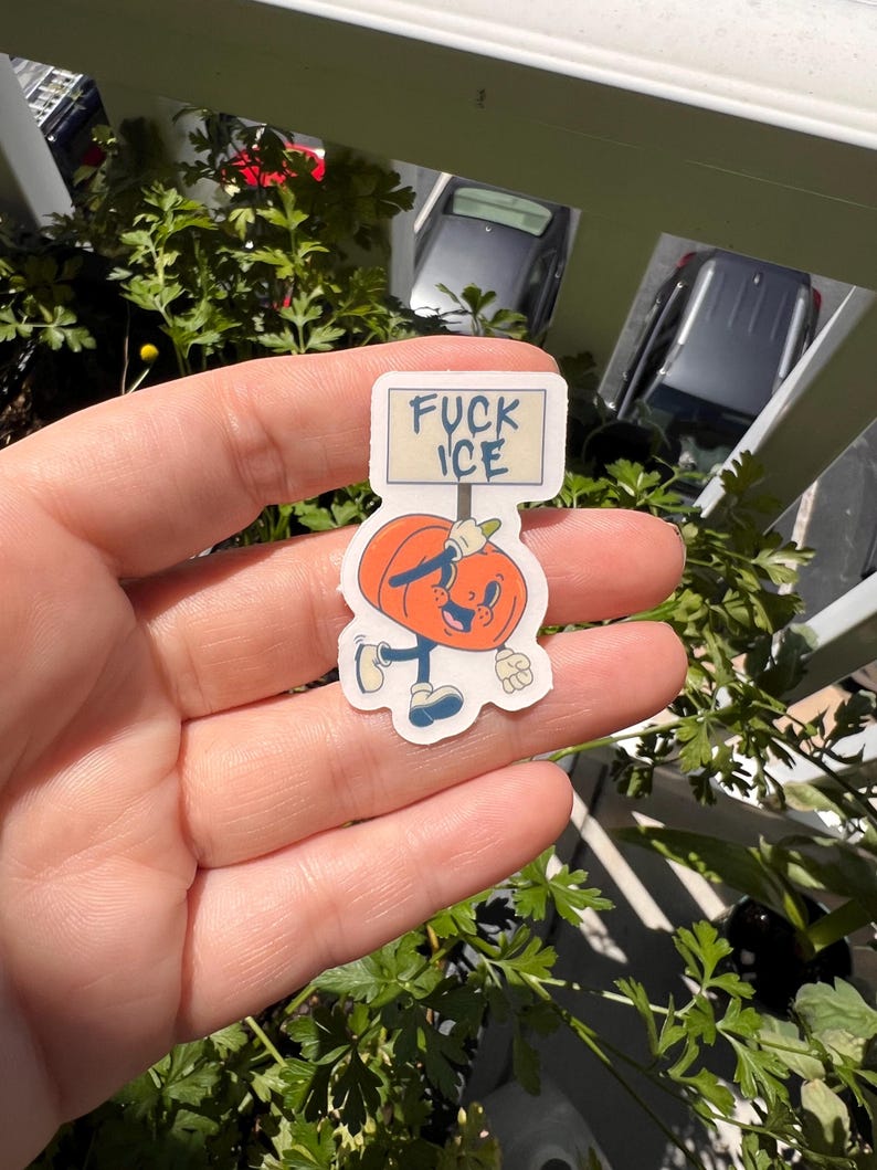 May include: A sticker featuring a cartoon pumpkin character holding a sign that reads "FUCK ICE". The pumpkin is orange with a smiling face, blue shoes, and a white glove. The sticker is held in a hand.