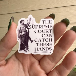 Op de afbeelding: Een witte sticker met een zwart-witte illustratie van de figuur van Justitia die een weegschaal en een zwaard vasthoudt. De tekst op de sticker luidt: "The Supreme Court Can Catch These Hands."