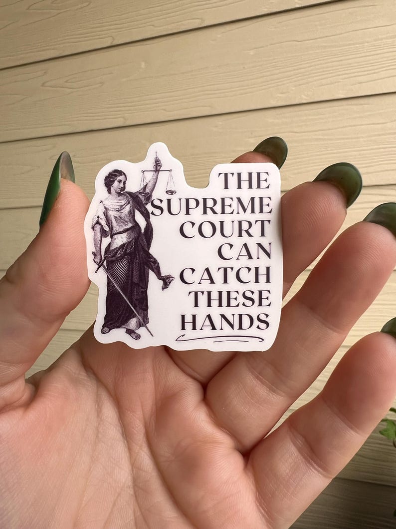 Op de afbeelding: Een witte sticker met een zwart-witte illustratie van de figuur van Justitia die een weegschaal en een zwaard vasthoudt. De tekst op de sticker luidt: "The Supreme Court Can Catch These Hands."
