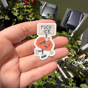 May include: A sticker featuring a cartoon pumpkin character holding a sign that reads "FUCK ICE". The pumpkin is orange with a smiling face, blue shoes, and a white glove. The sticker is held in a hand.