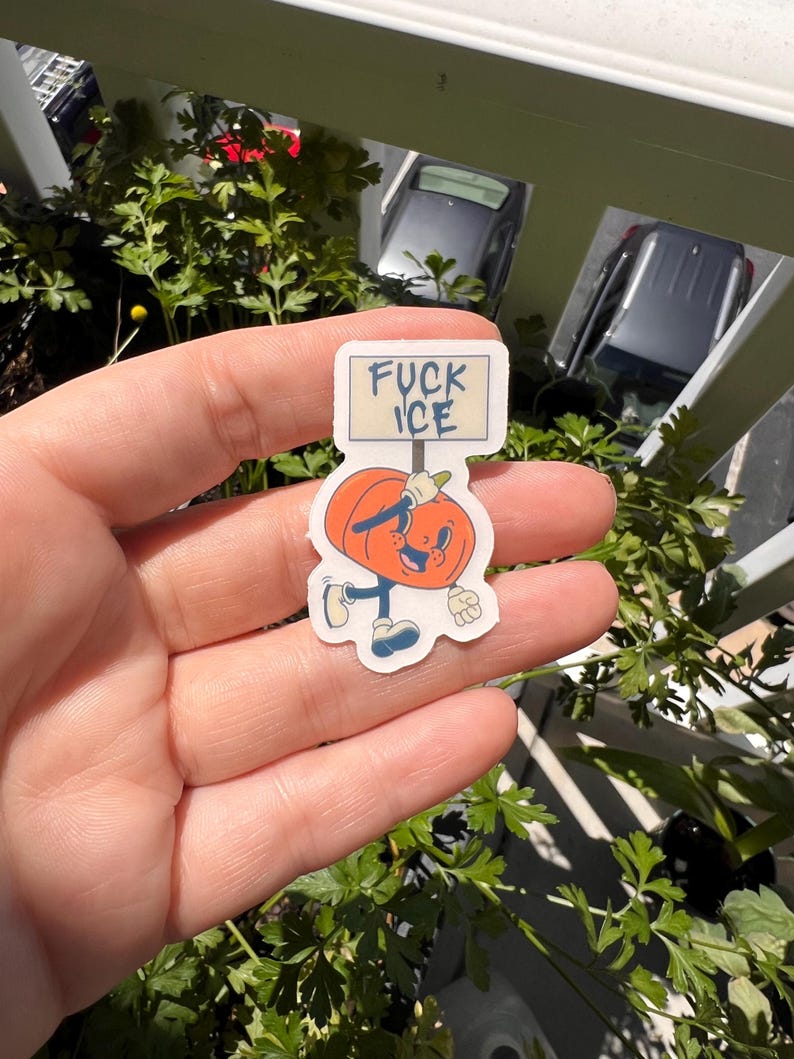 May include: A sticker featuring a cartoon pumpkin character holding a sign that reads "FUCK ICE". The pumpkin is orange with a smiling face, blue shoes, and a white glove. The sticker is held in a hand.