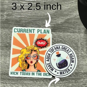 May include: Two sticker designs. The first is a 3 x 2.5 inch sticker with a retro comic book style illustration of a woman with blonde hair and large glasses. The text reads "CURRENT PLAN" and "KICK TODAY IN THE DICH". The second sticker is a 2 x 2 inch round sticker with a black background and white text that reads "MAKE AQUA TOFANA GREAT AGAIN" with a star design. The center of the sticker features a purple potion bottle with stars.