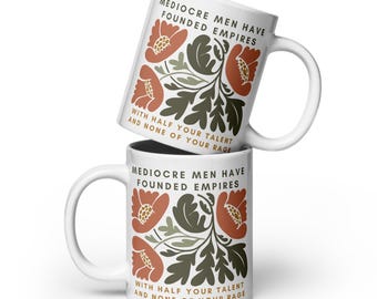 May include: Two white ceramic mugs stacked on top of each other. Each mug features a floral design with orange flowers and green leaves. The text on the mugs reads "MEDIOCRE MEN HAVE FOUNDED EMPIRES" and "WITH HALF YOUR TALENT AND NONE OF YOUR RAGE."