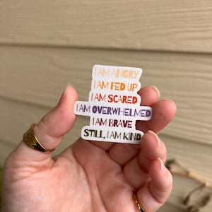 Könnte beinhalten: Ein weißer Aufkleber mit den Sätzen „I AM ANGRY“, „I AM FED UP“, „I AM SCARED“, „I AM OVERWHELMED“, „I AM BRAVE“ und „STILL, I AM KIND“ in verschiedenen Farben. Der Aufkleber wird in einer Hand mit goldenen Ringen gehalten.