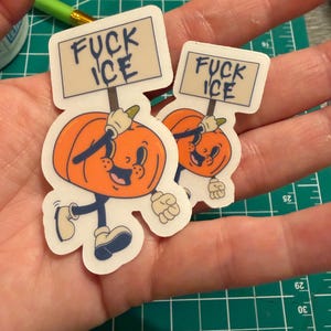 May include: Two die-cut vinyl stickers featuring a cartoon pumpkin character with a smiling face, wearing white gloves and boots, holding a rectangular sign that reads FUCK ICE in dark blue lettering against a light beige background.