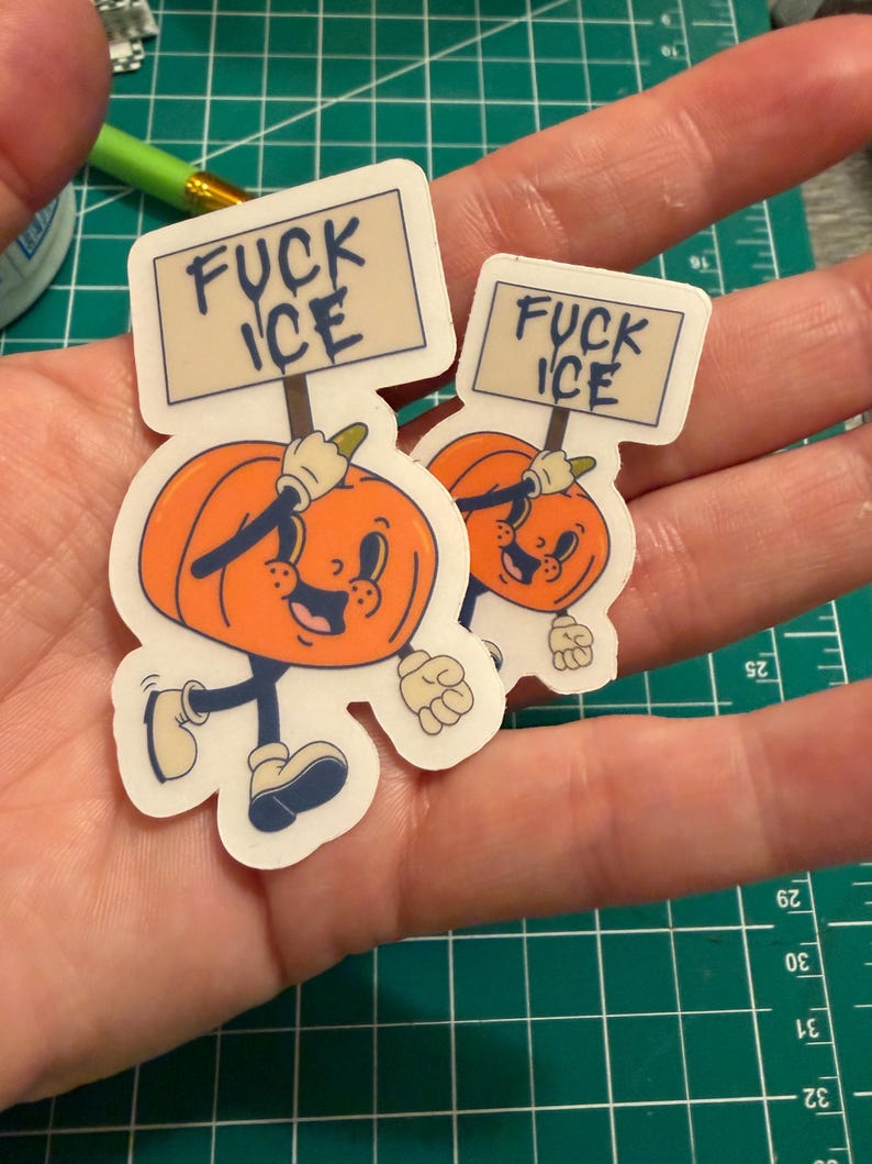 May include: Two die-cut vinyl stickers featuring a cartoon pumpkin character with a smiling face, wearing white gloves and boots, holding a rectangular sign that reads FUCK ICE in dark blue lettering against a light beige background.
