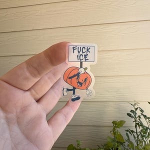 May include: A clear sticker featuring a cartoon pumpkin character holding a sign that reads "FUCK ICE". The pumpkin is orange with a smiling face, black shoes, and white gloves. The sticker is held up by a hand.