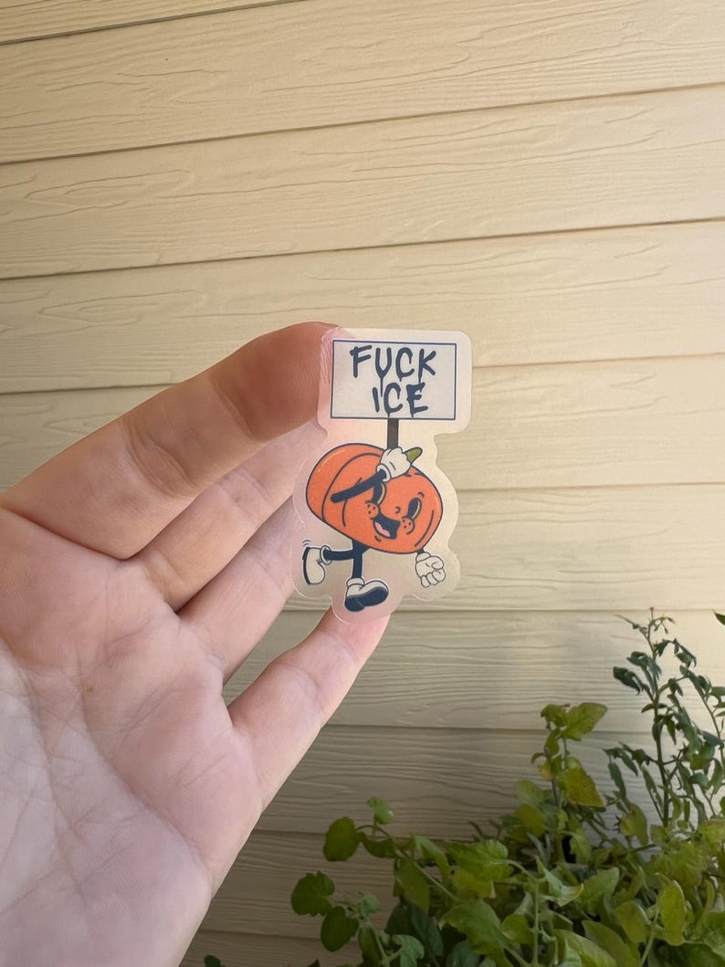 May include: A clear sticker featuring a cartoon pumpkin character holding a sign that reads "FUCK ICE". The pumpkin is orange with a smiling face, black shoes, and white gloves. The sticker is held up by a hand.