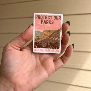 May include: A pink and brown sticker with the text "Protect Our Parks Not The Patriarchy" and a mountain scene.