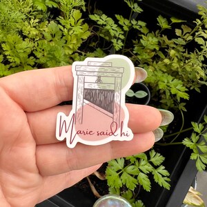 May include: A sticker with a drawing of a guillotine and the text "Marie said hi" in pink script. The sticker is on a hand with a green plant in the background.
