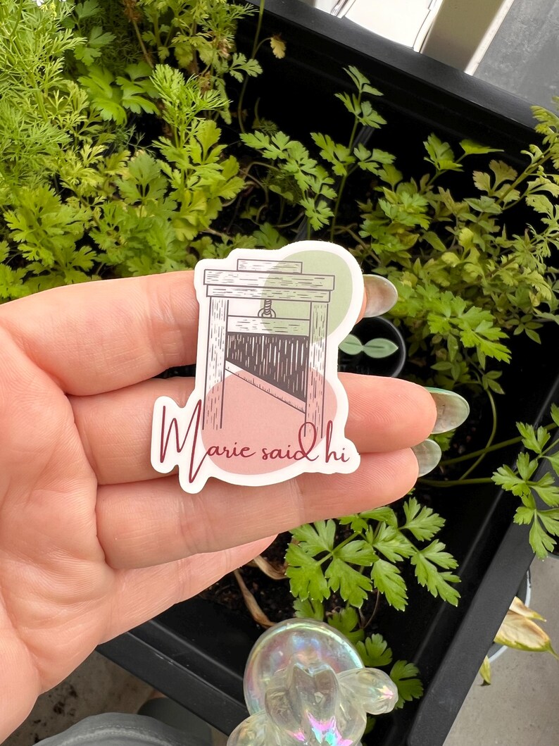 May include: A sticker with a drawing of a guillotine and the text "Marie said hi" in pink script. The sticker is on a hand with a green plant in the background.