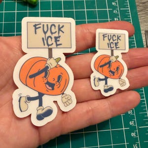 May include: Two die-cut vinyl stickers featuring a cartoon orange pumpkin character with a smiling face, wearing white gloves and blue shoes, holding a rectangular sign that reads FUCK ICE in dark blue lettering against a light background.