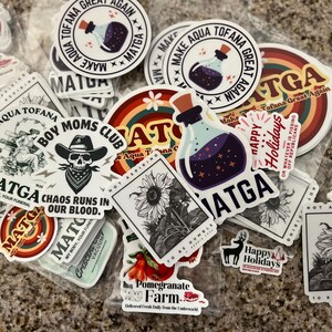 May include: A collection of colorful stickers featuring various designs, including a skull with a bandana, a sunflower, a bottle with a purple liquid, and the text "Make Aqua Tofana Great Again".