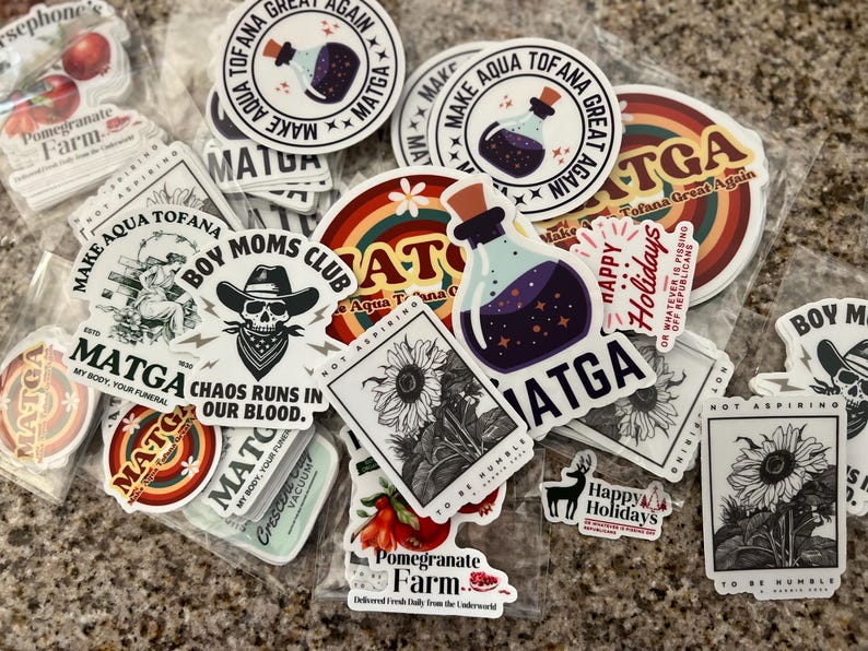 May include: A collection of colorful stickers featuring various designs, including a skull with a bandana, a sunflower, a bottle with a purple liquid, and the text "Make Aqua Tofana Great Again".