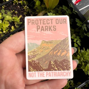 May include: A pink and white sticker with a mountain landscape and the text "Protect Our Parks Not The Patriarchy".