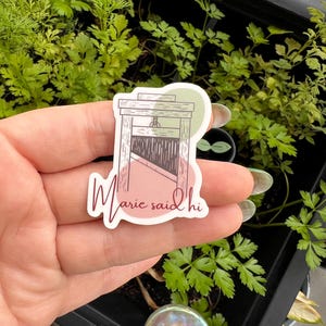 May include: A sticker with a pink and green background featuring a drawing of a guillotine with the text "Marie said hi" in cursive.