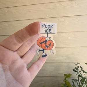 May include: A clear sticker featuring a cartoon pumpkin character holding a sign that reads "FUCK ICE." The pumpkin is orange with a smiling face, black shoes, and gloves. The sticker is held in a hand.