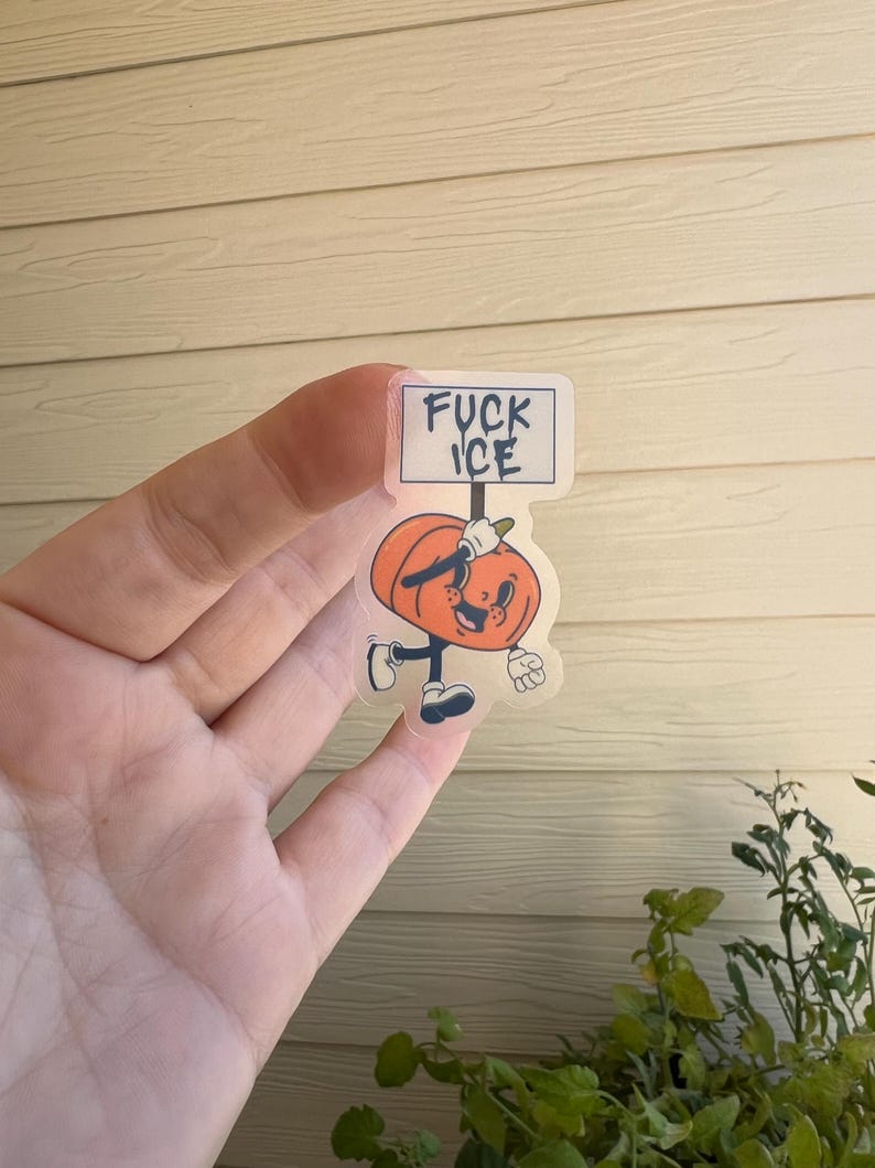 May include: A clear sticker featuring a cartoon pumpkin character holding a sign that reads "FUCK ICE." The pumpkin is orange with a smiling face, black shoes, and gloves. The sticker is held in a hand.