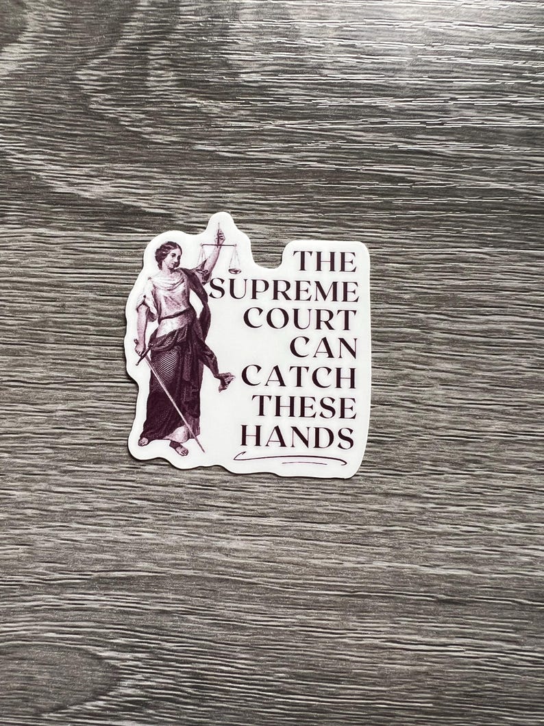 Op de afbeelding: Een witte sticker met een zwart-witte illustratie van een vrouw die een weegschaal en een zwaard vasthoudt. De tekst op de sticker luidt "The Supreme Court Can Catch These Hands".