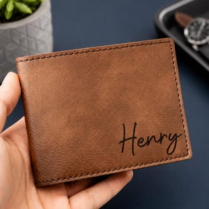 May include: A brown leather bifold wallet featuring a smooth, matte texture and dark brown stitching along the edges. The name Henry is embossed in a black, cursive script font on the bottom right corner of the front exterior of the wallet.
