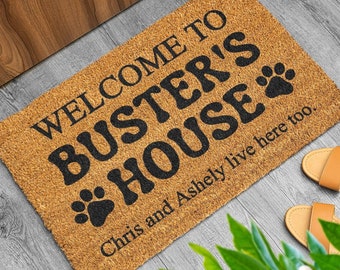 May include: Welcome to Buster's House doormat with a brown coir background and black text. The text reads "Welcome to Buster's House" with two paw prints below the word "House". The text "Chris and Ashley live here too." is printed below the paw prints.