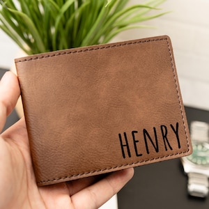 May include: Brown leather wallet with the name "HENRY" printed in black. The wallet is rectangular and has a stitched border. A watch with a silver band and green face is visible in the background.