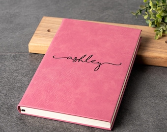 May include: A pink journal with the name "ashley" in black script. The journal is on a wooden cutting board, with a dark gray surface in the background. A small green plant is in the background.