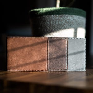 May include: Three rectangular bi-fold wallets arranged side-by-side. The first wallet is light brown, the second is a dark distressed brown, and the third is a solid gray. Each wallet features visible perimeter stitching and a smooth texture.