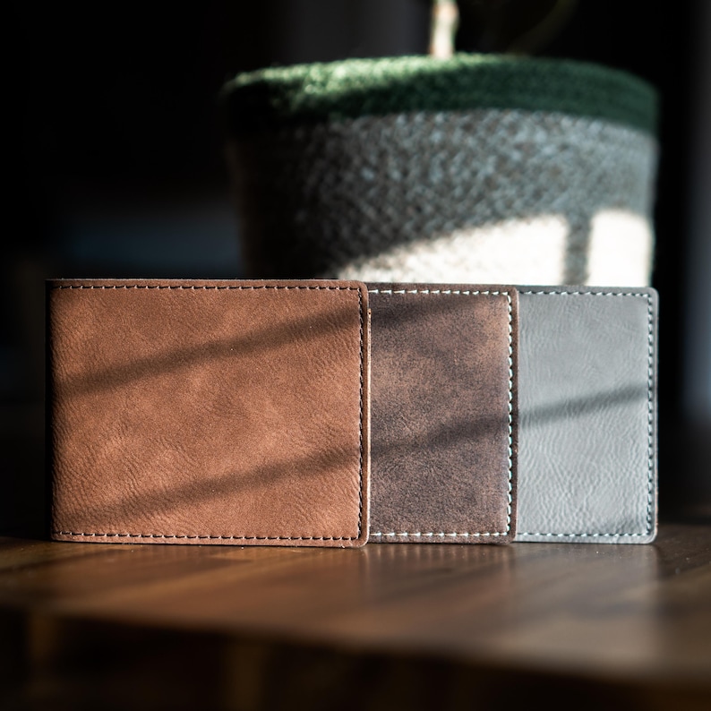 May include: Three rectangular bi-fold wallets arranged side-by-side. The first wallet is light brown, the second is a dark distressed brown, and the third is a solid gray. Each wallet features visible perimeter stitching and a smooth texture.