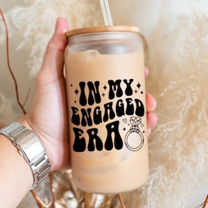 May include: A clear glass tumbler with a wooden lid and a straw, filled with a creamy beverage. The tumbler features the text "IN MY ENGAGED ERA" in black, along with a diamond ring graphic. A silver watch is visible.