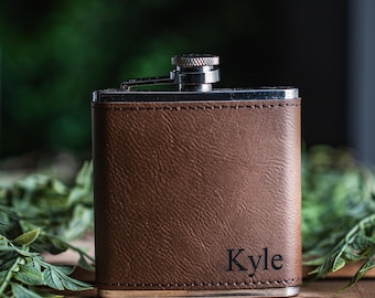 May include: A brown leather-wrapped flask with a silver cap and hinge. The flask has the name "Kyle" engraved on the front. The background is blurred, with green foliage and a wooden surface.