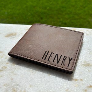 May include: A brown leather wallet with the name "HENRY" printed in black. The wallet is rectangular and appears to be folded. The background is blurred, with green grass visible.