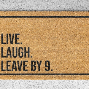 Op de afbeelding: Een bruine deurmat met zwarte tekst die "Live. Laugh. Leave by 9." luidt.