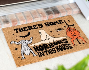 May include: A brown coir doormat with the text "There's some horrors in this house" in black lettering. The doormat features a cartoon mummy, ghost, and pumpkin with a spider hanging above.