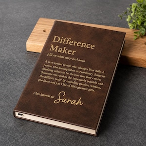 May include: A dark brown notebook with gold text on the cover. The text reads: Difference Maker [dif-er-uhns mey-ker] noun. A very special person who changes lives daily. Also known as: Sarah. The notebook has a smooth, matte-textured finish.