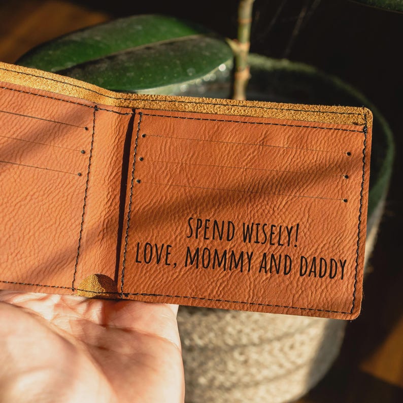 May include: A tan leather bifold wallet featuring black stitching and multiple card slots. The interior is engraved with the text: SPEND WISELY! LOVE, MOMMY AND DADDY. The wallet has a textured surface and a visible raw edge along the top fold.