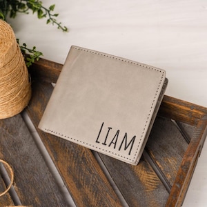May include: A light gray bi-fold wallet featuring the name LIAM embossed in a dark, sans-serif font on the bottom right corner. The wallet has visible tonal stitching along the edges and a smooth, matte finish on the exterior surface.