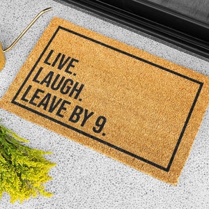 Op de afbeelding: Een bruine kokosvezel deurmat met zwarte tekst die "Live. Laugh. Leave by 9." luidt.