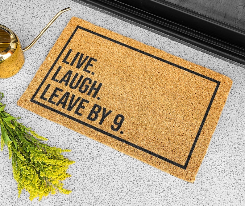 Op de afbeelding: Een bruine kokosvezel deurmat met zwarte tekst die "Live. Laugh. Leave by 9." luidt.
