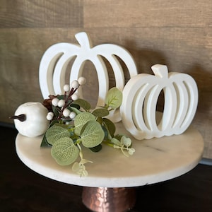 May include: White wooden pumpkin cutout decorations on a white marble cake stand with a copper base. The stand is decorated with a sprig of green eucalyptus and white berries.