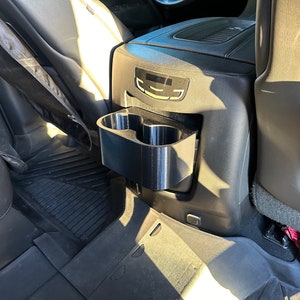 May include: Black plastic cup holder for a car. The cup holder is attached to the back of the front seat and has two cup slots.